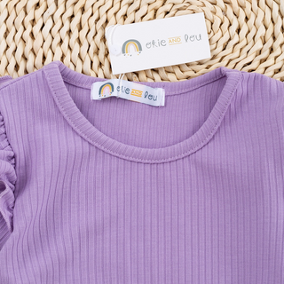 A close-up of a ribbed lavender purple children's top with a scoop neckline, laid flat on a woven straw mat. The brand tag 'etie AND deu' is visible on the collar and attached near the neckline.