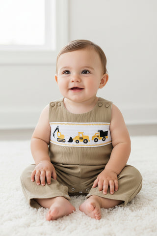 A baby's tan bib overalls romper with a white smocked chest panel featuring yellow construction vehicles (excavator, dump truck, and tow truck) and a yellow and white decorative border.