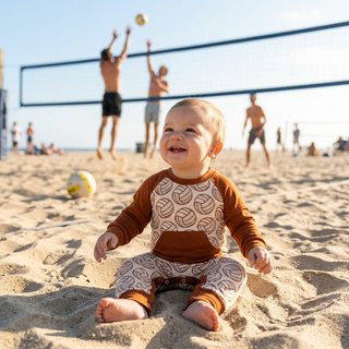 A baby sitting on sandy beach sand wearing a brown and cream patterned long-sleeve romper, with beach volleyball players visible in the background under a bright sky.
