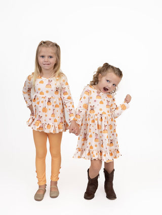 Two young girls standing and holding hands, wearing matching long-sleeve dresses with an orange and yellow autumn leaf pattern on a cream background, photographed against a solid white studio background.