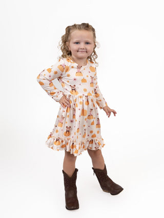 A young girl with curly blonde hair stands against a white studio background wearing a long-sleeve cream dress with an orange pumpkin and brown deer print, paired with dark brown cowboy boots.