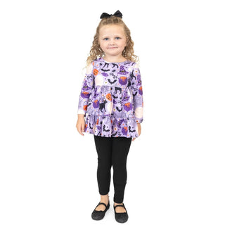 A young girl with curly blonde hair wearing a purple Halloween-themed long-sleeve ruffle top, black leggings, and black shoes, standing against a white background.