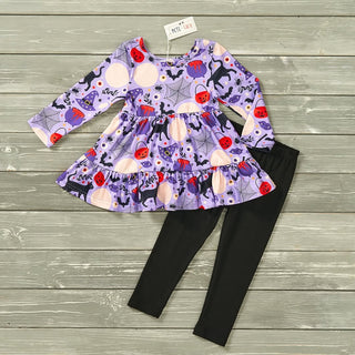 A toddler girl's Halloween-themed two-piece outfit featuring a purple long-sleeve ruffled top with a pumpkin, spider, bat, and basket print, paired with solid black leggings, laid flat on a gray wood plank background.