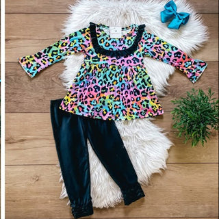 A children's outfit set displayed in a collage on a wood floor with a white faux-fur rug. The set includes a long-sleeve top and matching skirt in a bright multicolor leopard print, plus black leggings.