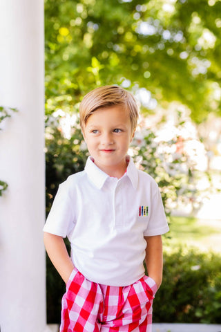 Young boy wearing a white polo shirt with a logo and red checkered shorts outdoors.