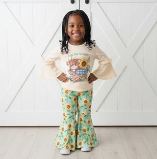 Child wearing a cream sweater with a sunflower design and green floral pants in front of a white wooden door.