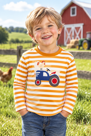A smiling young boy stands outdoors on a farm wearing a long-sleeve shirt with orange and white horizontal stripes and a tractor-and-pig graphic on the front, paired with blue jeans. A red barn, green fields, and a chicken are visible in the background.