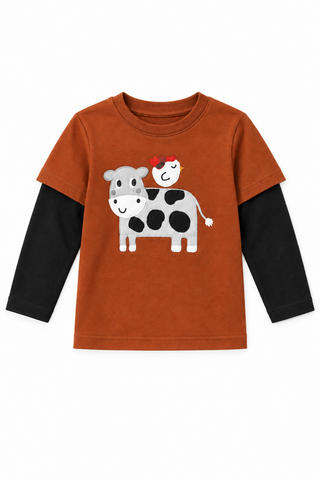 A young boy stands outdoors on a farm wearing a red long-sleeve shirt with a cow and pig graphic over a black long-sleeve layer, paired with blue jeans and brown boots.