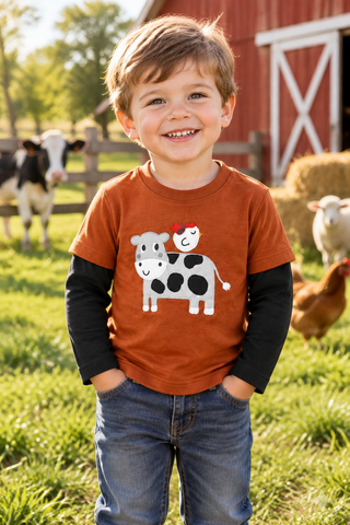 A young boy stands on a sunny farm wearing a rust-orange graphic t-shirt over a black long-sleeve shirt, paired with blue jeans. The t-shirt features a cow and sheep graphic.