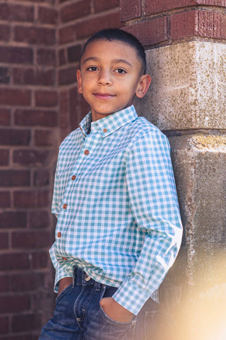 A young boy wearing a blue checkered shirt with a collar, paired with blue jeans, standing against a brick wall.
