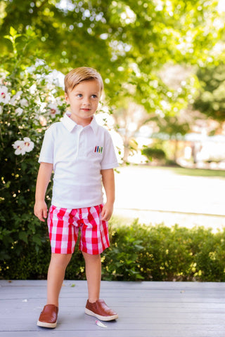 Young boy in a white polo shirt and red checkered shorts standing outdoors.
