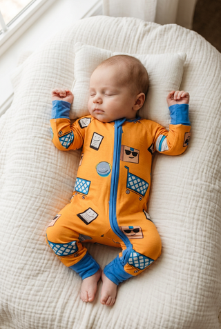 A sleeping baby wearing a bright orange zip-front onesie with blue cuffs and a shopping cart print, lying on a textured cream pillow.