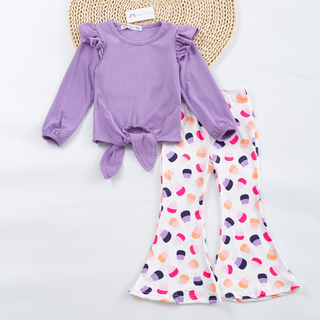 A two-piece girls' outfit set featuring a lavender ribbed long-sleeve top with ruffled shoulders and a front knot, paired with white bell-bottom pants covered in a colorful pastel semicircle pattern, displayed flat on a white background with a woven mat.