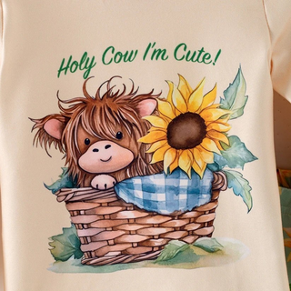 A close-up of a cream-colored garment featuring a watercolor-style graphic of a fluffy brown highland cow in a wicker basket next to a yellow sunflower, with the text 'Holy Cow I'm Cute!' printed above.
