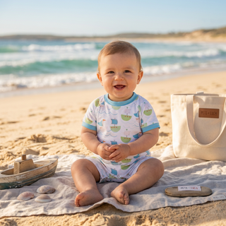 A baby sitting on a blanket on a sandy beach, wearing a white short-sleeve onesie with a blue and green boat pattern. A cream canvas tote bag with a brown 'Pitt Island' label sits to the right.