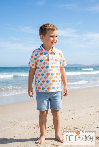 A young boy stands on a sandy beach wearing a short-sleeve polo shirt with a colorful fish pattern and light blue denim shorts, with the ocean and blue sky in the background.