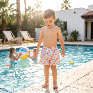 A young boy stands barefoot at the edge of a swimming pool wearing light pink swim trunks with a colorful bubble pattern. Two children are playing with a beach ball in the water behind him, with palm trees, lounge chairs, and a white stucco house in the background.