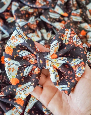 A close-up of a hair bow with an alligator clip, featuring a pattern of pumpkin spice and Liverpool fabric.