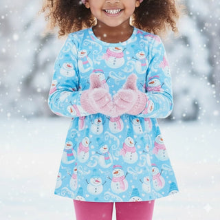 A young girl wearing a light blue long-sleeve dress with a snowman pattern, pink knit mittens, and pink leggings, standing in a snowy outdoor setting with falling snow.
